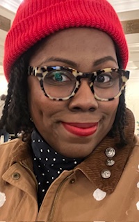 Kat Peace, portrait — wearing a red beanie, tortoiseshell glasses, and a tan jacket.